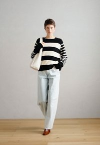 Pull de riscas preto e branco, jeans largos azul claro e sapatos de salto castanho; modelo posa com uma mala de ombro branca contra um fundo simples.