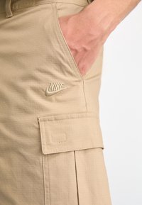 Vaaleat Nike cargo-housut, joissa on käsi sivutaskussa, näyttäen brodeeratun logon ja kankaan rakenteen.