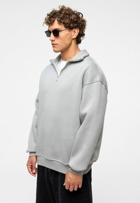 Man met krullend haar die zwarte zonnebril draagt, een lichtgrijze half-zip sweatshirt, een witte onderhemd en zwarte broek tegen een neutrale achtergrond.