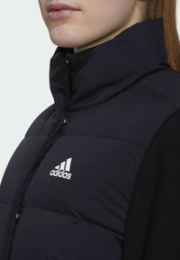Zwarte puffer vest met hoge kraag, drukknoopsluiting en wit Adidas-logo aan de linkerkant, met een gladde, matte textuur.