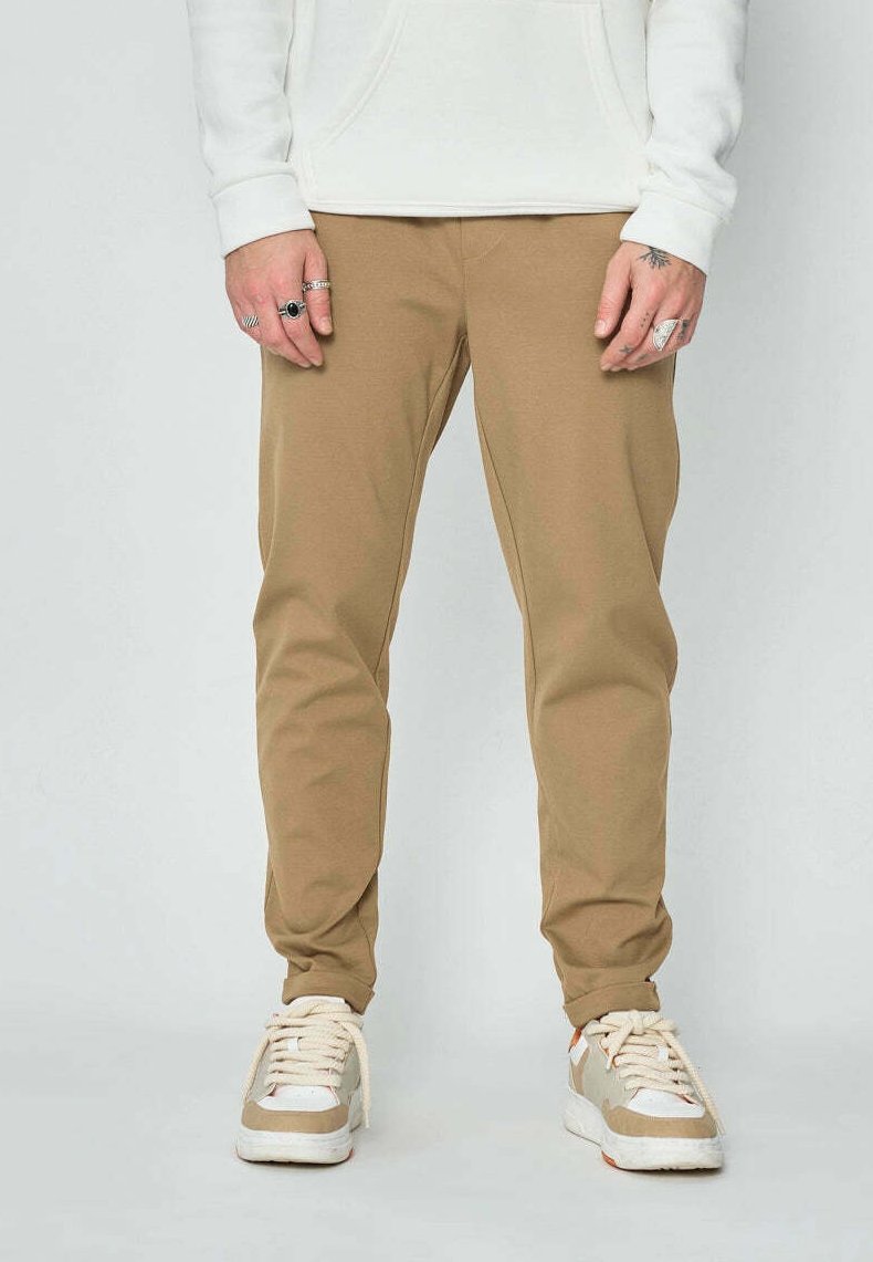 Homme portant un pantalon beige, un sweat-shirt blanc et des baskets beige et blanches, se tenant avec les mains détendues le long du corps contre un fond uni.