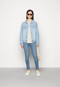 Veste en denim bleu clair avec poches, haut beige, skinny jeans bleu clair et baskets crème. Accessoirisée avec de petites lunettes de soleil noires et un sac beige.