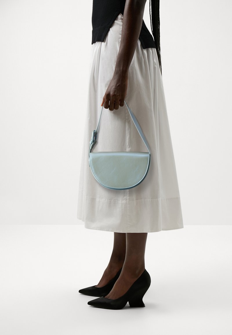 Femme portant une jupe blanche et un haut noir tient un sac à main bleu clair en forme de demi-lune, chaussée de talons noirs sur un fond uni.