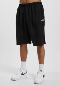 Sorte bomulds shorts med elastisk talje, der har trykt hvid tekst "DEF" på venstre ben. Båret med hvide sneakers og strømper.