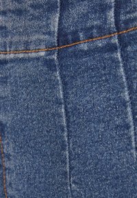 Sininen denim-kangas, jossa näkyy oranssia ompelua ja haalistunut tekstuuri sauman varrella, osoittaen yksityiskohtaista kudosta ja kulumakuvioa.