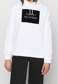 Felpa bianca con cappuccio e logo rettangolare nero con la scritta "TOMMY HILFIGER". Maniche lunghe e cordini sul cappuccio. Tessuto morbido.