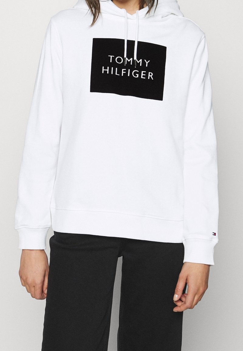 Felpa bianca con cappuccio e logo rettangolare nero con la scritta "TOMMY HILFIGER". Maniche lunghe e cordini sul cappuccio. Tessuto morbido.