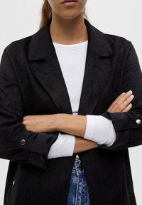 Black faux-suede blazer with a notched collar, long sleeves, and button accents, layered over a white fitted top.