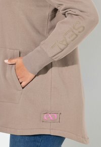 Mano nella tasca di una felpa beige con la scritta "SEAL" ricamata sul manica e una patch rosa a forma di infinito vicino all'orlo.