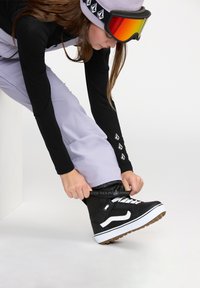 Bottes de neige noires avec des accents blancs, design à lacets, semelle en caoutchouc, matériau texturé. Une personne ajuste les bottes tout en portant un pantalon violet clair.