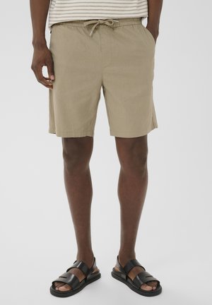 Mand iført beige shorts med snøre, stribet skjorte og sorte lædersandaler, stående med hænderne i lommerne.