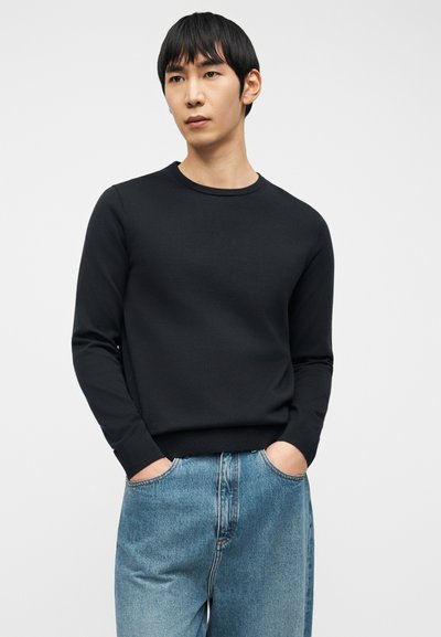 Άνδρας με μαύρα μαλλιά που φοράει μαύρο μακρυμάνικο πουλόβερ με στρογγυλή λαιμόκοψη και μπλε τζιν, στέκεται με τα χέρια στις τσέπες μπροστά σε φωτεινό φόντο.