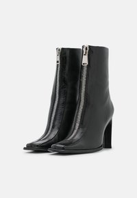 Paire de bottines en cuir noir élégantes à talons hauts, avec des fermetures éclair argentées à l'avant, des bouts pointus et des talons empilés sur fond blanc.