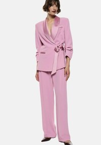 Completo rosa con giacca lunga a scollatura profonda a V, dettagli in satin e pantaloni a gamba larga con una consistenza morbida. Cintura in vita.