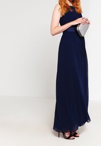 Kvinna i en golvlång marinblå klänning, som håller en silverclutch och har på sig svarta högklackade sandaler, stående mot en enkel vit bakgrund.