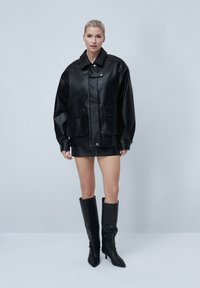 Veste noire en cuir courte avec deux poches avant, large col et fermeture à boutons, portée sur une jupe noire et des bottes montantes.