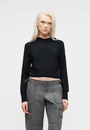 Młoda osoba o długich blond włosach, ubrana w czarny krótki sweter i szare spodnie typu cargo, stojąca na jednolitym jasnym tle.