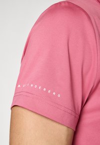 Camisa rosa de mangas curtas feita de tecido liso. Possui um logótipo branco na manga esquerda e um design limpo e moderno.