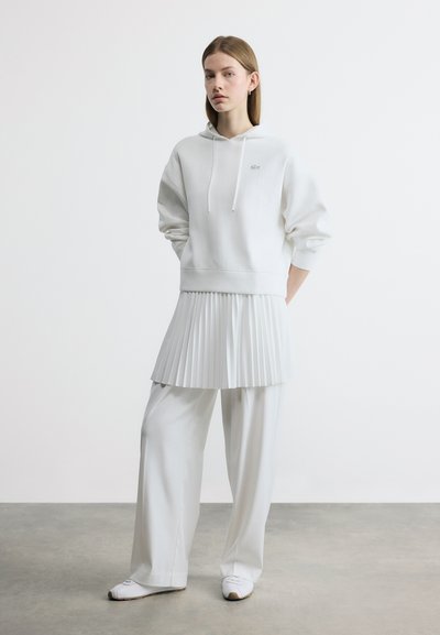Jeune femme debout portant un sweat à capuche blanc surdimensionné, une mini-jupe plissée par-dessus un pantalon large, et des baskets blanches sur un fond uni.