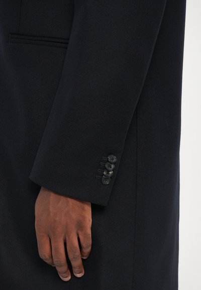 Manica di un cappotto blu navy con una finitura testurizzata, caratterizzata da quattro bottoni neri sul polsino e un tessuto liscio visibile sopra il polso.