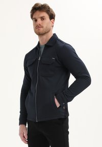 Chaqueta de naví con cremallera, dos bolsillos en el pecho, mangas largas y cuello. Fabricada con un tejido texturizado y un diseño ajustado.