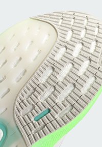 Schuhsohle mit einer strukturierten weißen Gummibasis, die transparente Abschnitte und grüne Akzente aufweist. Besonders auffällig ist das dreidimensionale Grip-Design und die Aussparungen.