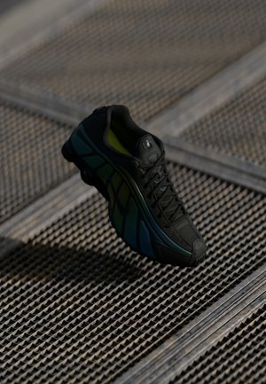 Chaussure de sport noire avec des touches bleues et vertes suspendue au-dessus d’un sol en grille métallique texturée dans une faible luminosité.