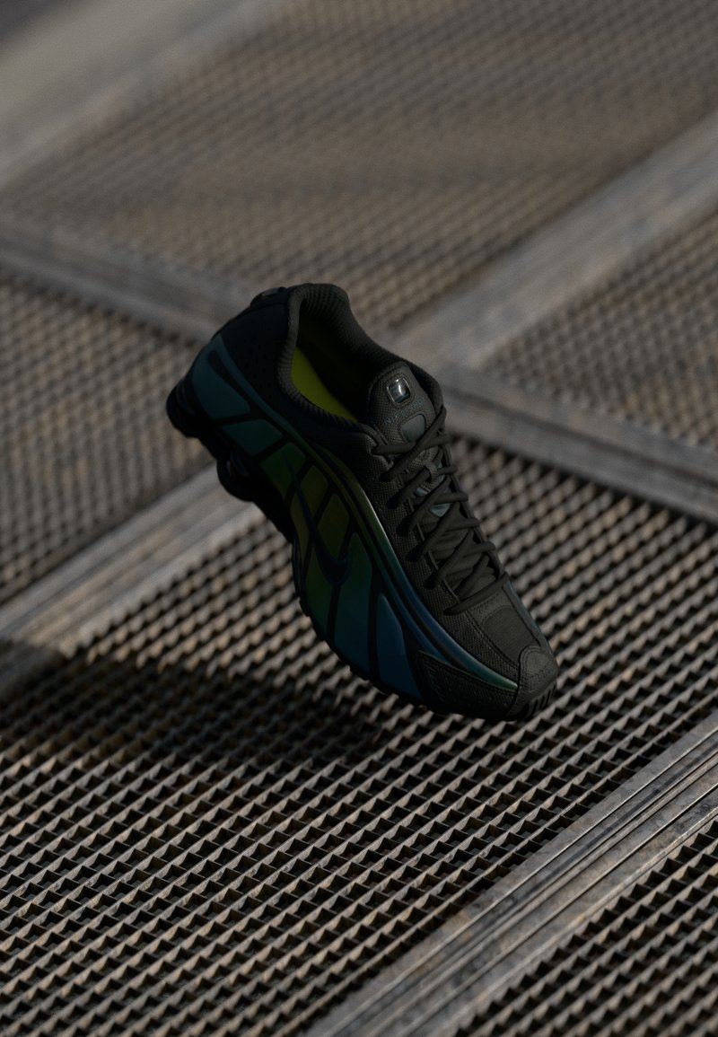 Chaussure de sport noire avec des touches bleues et vertes suspendue au-dessus d’un sol en grille métallique texturée dans une faible luminosité.
