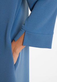 Hand in der Tasche eines blauen Kleidungsstücks mit strukturierter Stoffoberfläche und weit geschnittenem, cuffed Ärmel mit einem Schlitz auf der Vorderseite.