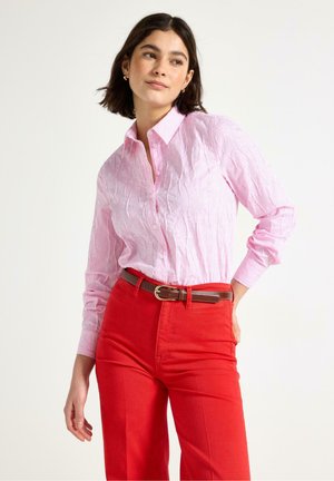 Donna con camicia a righe rosa chiaro con bottoni infilata in pantaloni rossi a vita alta con cintura marrone, in piedi con una mano sul fianco.
