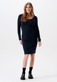 Mörkblå långärmad klänning med omslagdesign och rynkad sidodetalj, tillverkad av ett stretchnära tyg. Svarta platåskor och strumpor som når knäna.