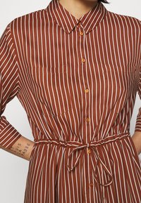 Robe chemise rayée marron et blanche avec un col, une fermeture à boutons sur le devant, deux poches poitrine et une ceinture pour l'ajustement.