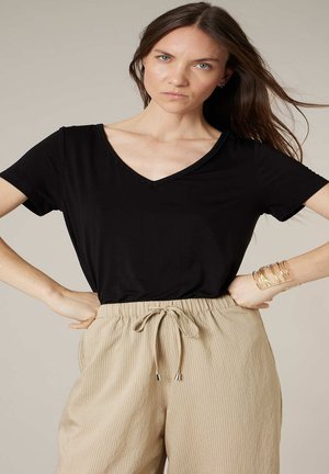 Vrouw met lang bruin haar, die een zwart V-hals shirt en beige broek met trekkoord draagt, staand met handen in de zij tegen een neutrale achtergrond.