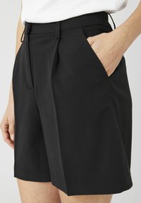 Shorts noirs sur mesure avec un devant plat, deux poches latérales et une texture lisse, présentant des plis pour plus de structure et de forme.