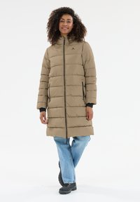 Beige puffervest med vandret syninger, høj krave, to sidelommer og lynlås, båret over lyseblå jeans og sorte støvler.