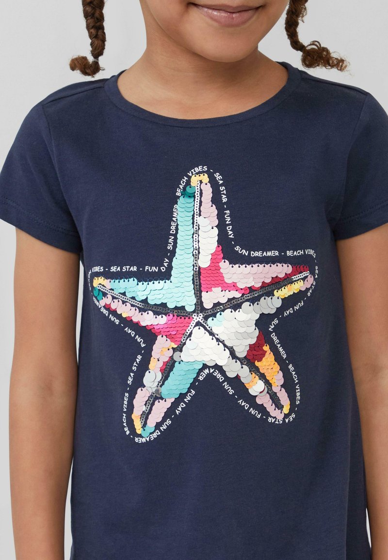 Marineblauwe t-shirt met een kleurrijke zeesterafbeelding gemaakt van glanzende pailletten, met tekst die het ontwerp in een cirkelvormige indeling omringt.