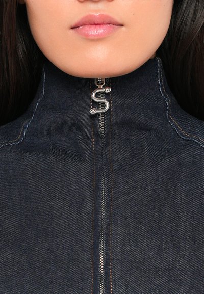 Temno modra denim jakna z visokim ovratnikom, z srebrnim zadrgo in akcentom v obliki črke "S" ter kontrastnim oranžnim šivanjem.