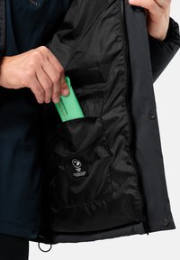 Veste isolante noire avec une fermeture éclair à l'avant. Une main sort un objet vert d'une poche intérieure. Présence d'une étiquette en tissu sur la doublure.