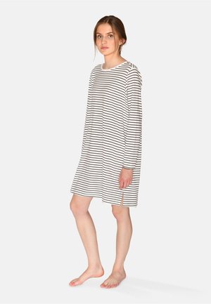 Junge Frau steht barfuß und trägt ein langärmliges, weißes Kleid mit schwarzen horizontalen Streifen und seitlichem Schlitz.