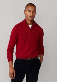 Roter Reißverschluss-Pullover mit gerippten Bündchen und Saum, aus weichem Material, kombiniert mit schwarzen Hosen und einem braunen Gürtel.