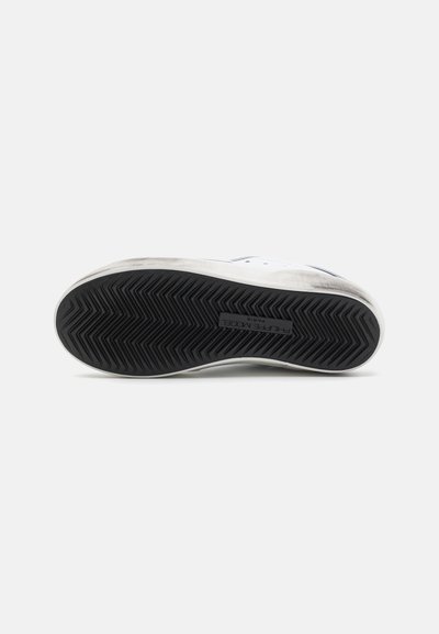 Fehér sneaker, texturált fekete gumitalppal, amelyen zig-zag mintázat és finom márkajelzés található a szegélyen.