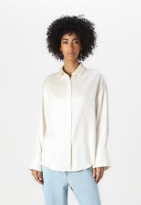 Kremfarget silkebluse med knappefront, spiss krage og lange ermer med brede mansjetter. Brukt med lyseblå jeans.