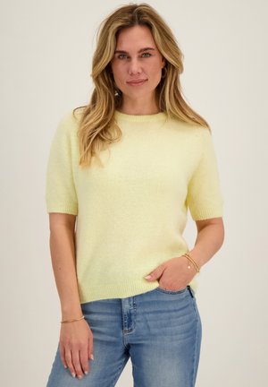 Vrouw met lang blond haar die een lichtgeel kort mouwtruitje en een blauwe spijkerbroek draagt, staand met één hand in de broekzak en een neutrale achtergrond.