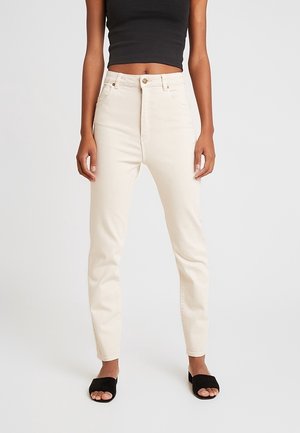 Mujer con vaqueros de tiro alto color blanco roto, top negro corto y sandalias negras de tiras abiertas, de pie sobre un fondo liso.