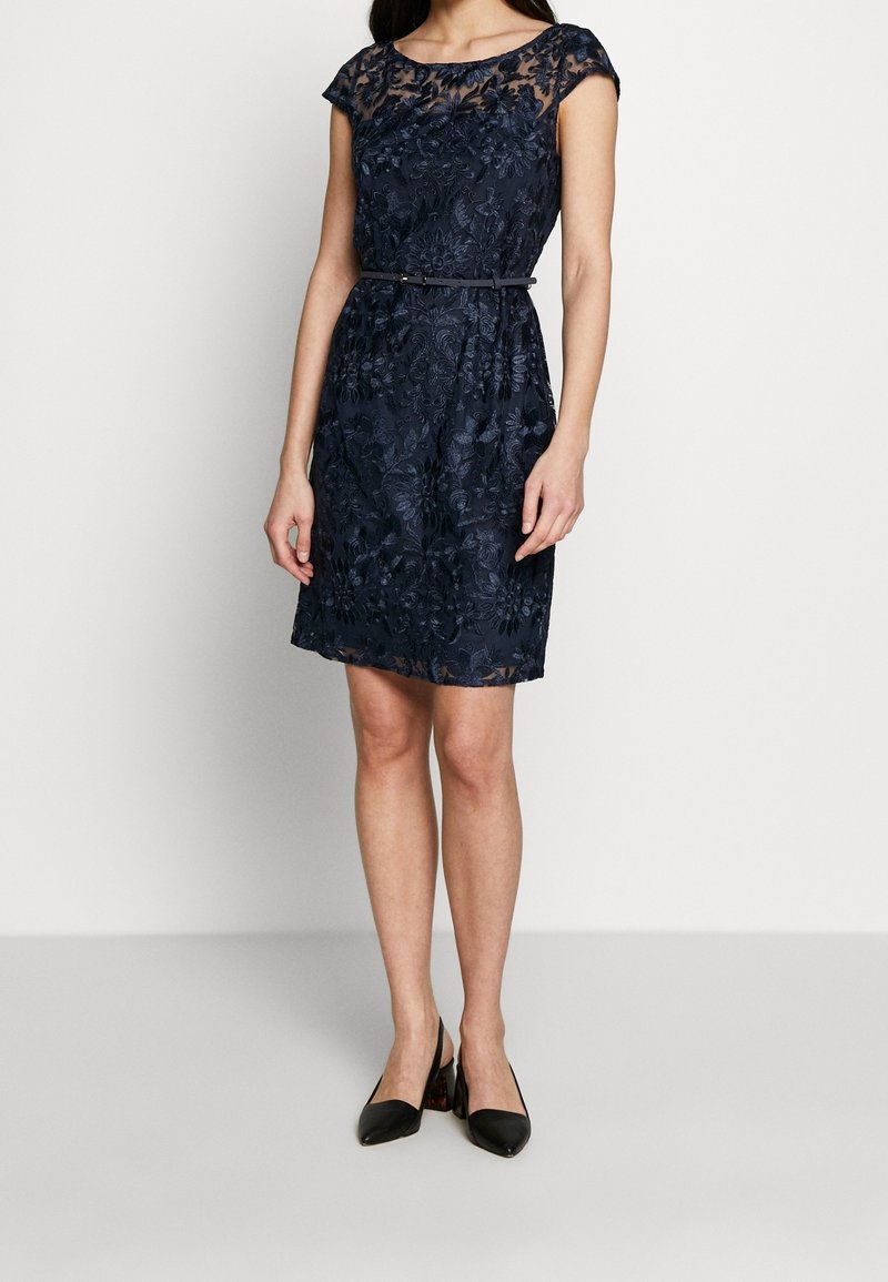 Une robe en dentelle bleu marine présente un décolleté en vagues, des manches courtes, des motifs floraux et une taille cintrée avec une ceinture assortie. Des talons noirs.