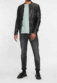 Černá kožená bunda s vysokým límcem, zelené tričko pod ní, šedé slim-fit džíny a černé boty s tlustou podrážkou.