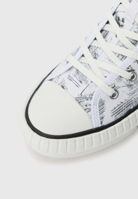 Hvite sneakers med et teksturert, mønstret canvas-overdel som har svarte grafiske design. Gummitåhette og bølget hvit såle. Snøring.