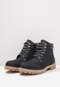 Botas de tobillo de ante azul oscuro con detalles en piel negra, ojales metálicos y una suela de goma resistente. Presentan cordones grises y un cuello acolchado.