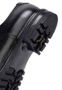 Bota de cuero negra con suela de goma gruesa y con tacos, que presenta patrones de huella geométricos profundos para una mayor tracción y durabilidad.
