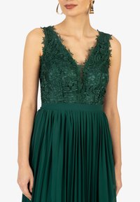 Grünes Abendkleid mit einem Spitzenoberteil mit Blumenmustern, einem tiefen V-Ausschnitt und einem plissierten Rock für Textur und Bewegungsfluss.